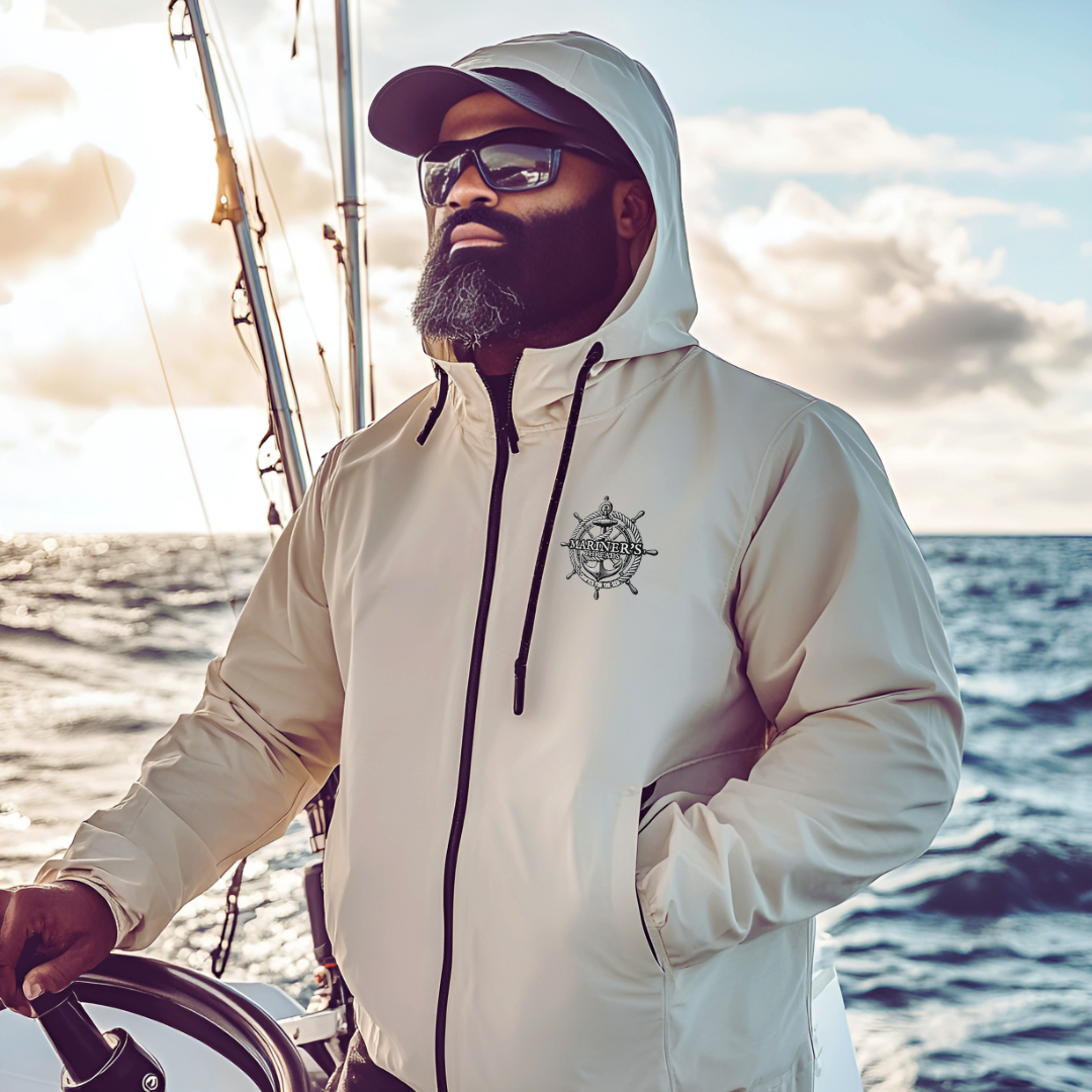 Man steering a boat on the open ocean, wearing a sleek beige Mariner’s Threads™ windbreaker with a black nautical helm logo on the chest. His hood is up, and he sports sunglasses and a beard, embodying the resilience and adventure of life at sea.